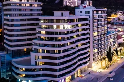 a tall white building at night with a city at Azure Coast Residence Rafailovici -Seaside Living Apartments in Becici