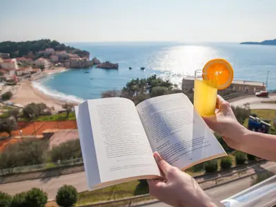 a person holding a book and a glass of orange juice at Apartments Raymond in Sveti Stefan