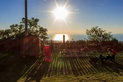 a man sitting on a bench in front of a fence with the sun at Old stone house with swimming pool Etno House Perazic in Bar