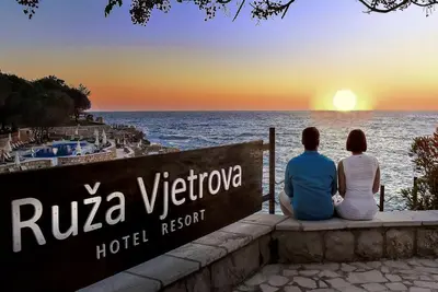 two people sitting on a sign looking at the ocean at Radisson Resort Ruza Vjetrova in Dobra Voda